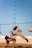 A person is playing beach volleyball, preparing to receive or bump the ball. The individual is wearing a sleeveless top and shorts, with a volleyball flying toward them. The background includes a blue sky and a net above a sand court.