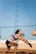 A person is playing beach volleyball, preparing to receive or bump the ball. The individual is wearing a sleeveless top and shorts, with a volleyball flying toward them. The background includes a blue sky and a net above a sand court.