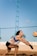 A person is playing beach volleyball, preparing to receive or bump the ball. The individual is wearing a sleeveless top and shorts, with a volleyball flying toward them. The background includes a blue sky and a net above a sand court.