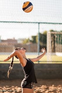 A woman is poised to serve a volleyball while standing on a sandy court. She has her arms stretched back, and her eyes are focused upwards at the ball in mid-air. Her hair is tied in a braid, and she is wearing athletic gear, including a tank top and shorts. In the background, there is a blurred view of a net, trees, and buildings, suggesting an outdoor setting.