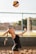 A woman is poised to serve a volleyball while standing on a sandy court. She has her arms stretched back, and her eyes are focused upwards at the ball in mid-air. Her hair is tied in a braid, and she is wearing athletic gear, including a tank top and shorts. In the background, there is a blurred view of a net, trees, and buildings, suggesting an outdoor setting.