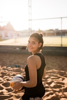 A young woman is standing on a sandy outdoor volleyball court, smiling and holding a volleyball under one arm. She is wearing a black sleeveless sports top and has her hair tied back. The background shows a net and some out-of-focus buildings under a blue sky with sunlight creating a warm, backlit effect.