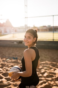 A young woman is standing on a sandy outdoor volleyball court, smiling and holding a volleyball under one arm. She is wearing a black sleeveless sports top and has her hair tied back. The background shows a net and some out-of-focus buildings under a blue sky with sunlight creating a warm, backlit effect.