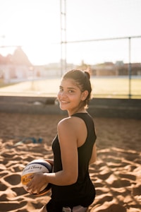 A young woman is standing on a sandy outdoor volleyball court, smiling and holding a volleyball under one arm. She is wearing a black sleeveless sports top and has her hair tied back. The background shows a net and some out-of-focus buildings under a blue sky with sunlight creating a warm, backlit effect.
