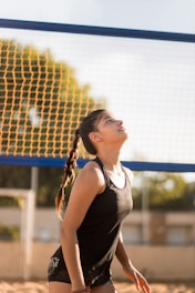 A young woman dressed in athletic wear stands on a sandy volleyball court, gazing upwards with focus. The volleyball net behind her has blue and yellow colors, with blurred green foliage in the background, indicating an outdoor setting.