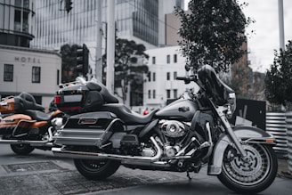 An urban setting with two parked motorcycles on the street. The foreground features a black touring motorcycle with chrome detailing and a large windscreen. The background includes a modern cityscape with buildings, trees, and urban fixtures creating a lively backdrop.