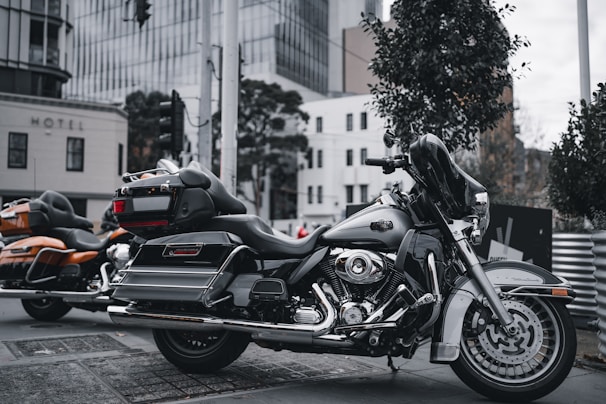 An urban setting with two parked motorcycles on the street. The foreground features a black touring motorcycle with chrome detailing and a large windscreen. The background includes a modern cityscape with buildings, trees, and urban fixtures creating a lively backdrop.