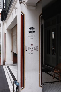 A white corner of a building featuring signage for Tianjing Hotel and Lim Kee Cafe, with decorative red and gold stripes. There's a wooden chair and tiled floor visible inside.