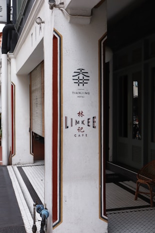 A white corner of a building featuring signage for Tianjing Hotel and Lim Kee Cafe, with decorative red and gold stripes. There's a wooden chair and tiled floor visible inside.