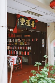 An entrance to a traditional Chinese shop featuring rows of colorful jars and containers filled with various goods. A sign with Chinese characters hangs above the doorway, adorned with red decorations. A tricycle cart with a fabric cover is parked outside the shop. The floor is tiled in a deep red, and green plants are visible in the foreground.