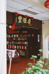 An entrance to a traditional Chinese shop featuring rows of colorful jars and containers filled with various goods. A sign with Chinese characters hangs above the doorway, adorned with red decorations. A tricycle cart with a fabric cover is parked outside the shop. The floor is tiled in a deep red, and green plants are visible in the foreground.