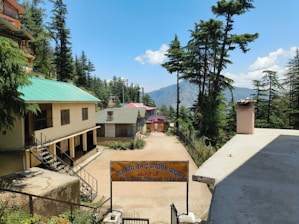 A rural school courtyard with a few buildings surrounded by tall pine trees and distant mountains under a clear blue sky. The main building has a beige facade with a green roof, accessed by a narrow staircase with black railings. A signboard in front is written in Hindi.
