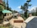 A rural school courtyard with a few buildings surrounded by tall pine trees and distant mountains under a clear blue sky. The main building has a beige facade with a green roof, accessed by a narrow staircase with black railings. A signboard in front is written in Hindi.