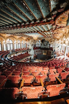An opulent theater interior with ornate architectural details, featuring a grand, colorful ceiling adorned with intricate patterns and sculptures. Rows of plush red seats fill the space, leading down to a stage area. Large arched windows allow natural light to illuminate the richly decorated environment.