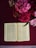 An open book lies on a richly textured maroon cloth, surrounded by a vibrant bouquet of pink and red flowers. The pages of the book display dense text in a clean, serif font. The scene is softly lit, highlighting the delicate details of the flowers and the intricate patterns of the cloth beneath.
