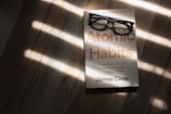 A pair of black rectangular eyeglasses rest on a book titled 'Atomic Habits' by James Clear. The book is placed on a wooden surface, with sunlight streaming through blinds, casting striped shadows across the scene.