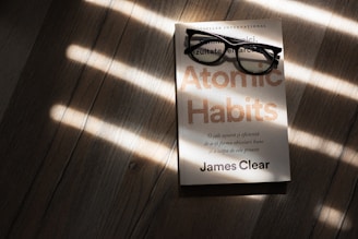 A pair of black rectangular eyeglasses rest on a book titled 'Atomic Habits' by James Clear. The book is placed on a wooden surface, with sunlight streaming through blinds, casting striped shadows across the scene.