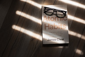 A pair of black rectangular eyeglasses rest on a book titled 'Atomic Habits' by James Clear. The book is placed on a wooden surface, with sunlight streaming through blinds, casting striped shadows across the scene.