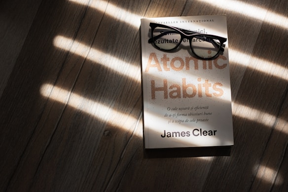 A pair of black rectangular eyeglasses rest on a book titled 'Atomic Habits' by James Clear. The book is placed on a wooden surface, with sunlight streaming through blinds, casting striped shadows across the scene.