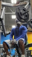 A person wearing a blue long-sleeve shirt, white and blue shorts, white sneakers, gloves, and a cap is sitting on a bench in front of a weight machine loaded with plates in a gym. The background shows gym equipment and bright wall colors in blue and yellow.