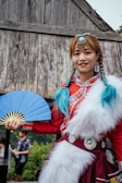 A person wearing traditional attire featuring a red dress with elaborate patterns and white fur details stands in front of a rustic wooden structure. They hold a blue fan in one hand and have braided hair adorned with blue accents and decorative jewelry on their head.