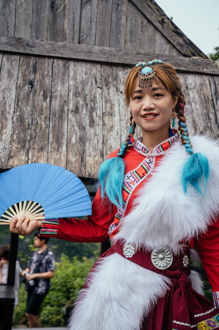 A person wearing traditional attire featuring a red dress with elaborate patterns and white fur details stands in front of a rustic wooden structure. They hold a blue fan in one hand and have braided hair adorned with blue accents and decorative jewelry on their head.