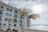 A modern, white multi-story building with several balconies facing a large body of water. Tall palm trees are planted near the building, with a bright, overcast sky in the background.