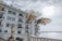A modern, white multi-story building with several balconies facing a large body of water. Tall palm trees are planted near the building, with a bright, overcast sky in the background.