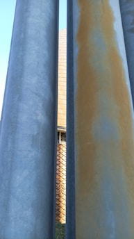 Several large cylindrical concrete pillars are positioned closely together in the foreground. Behind them, a textured brick wall with a geometric design is partially visible. The color gradient on the pillars varies from light blue to grey and rusty brown.