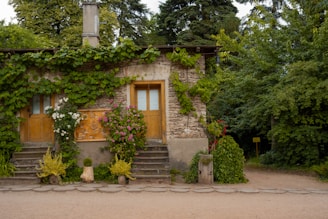 A rustic stone house with wooden doors surrounded by lush greenery and flowering plants. Ivy and vines cover the facade, adding to the natural aesthetic. The setting is tranquil, with mature trees in the background.