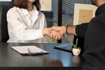 Two people sitting at a desk are shaking hands over paperwork, likely indicating the conclusion of a business deal or an agreement. One person is wearing a white shirt and the other is dressed in dark clothing. Other items present include a laptop, a small potted plant, and a clipboard with a pen.