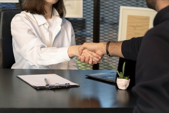 Two people sitting at a desk are shaking hands over paperwork, likely indicating the conclusion of a business deal or an agreement. One person is wearing a white shirt and the other is dressed in dark clothing. Other items present include a laptop, a small potted plant, and a clipboard with a pen.