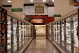 A grocery store aisle featuring glass-door freezers filled with various frozen goods. Signs above indicate sections for frozen entrees, pizzas, seafood, meats, breads, and other goods. Bright fluorescent lighting illuminates the area, highlighting different food packaging.