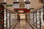 A grocery store aisle featuring glass-door freezers filled with various frozen goods. Signs above indicate sections for frozen entrees, pizzas, seafood, meats, breads, and other goods. Bright fluorescent lighting illuminates the area, highlighting different food packaging.