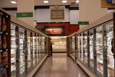 A grocery store aisle featuring glass-door freezers filled with various frozen goods. Signs above indicate sections for frozen entrees, pizzas, seafood, meats, breads, and other goods. Bright fluorescent lighting illuminates the area, highlighting different food packaging.