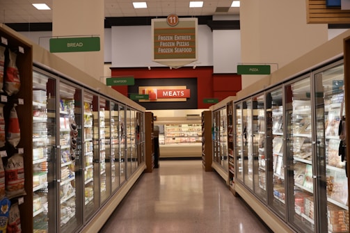 A grocery store aisle featuring glass-door freezers filled with various frozen goods. Signs above indicate sections for frozen entrees, pizzas, seafood, meats, breads, and other goods. Bright fluorescent lighting illuminates the area, highlighting different food packaging.