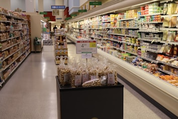 A grocery store aisle displaying various packaged foods on shelves. In the center, there is a promotional display of bags containing toffee popcorn, with a sign indicating the price. The aisle is well-lit, and the shelves are stocked with an assortment of products, including snacks, dairy, and bread.