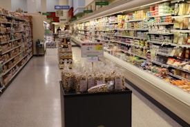 A grocery store aisle displaying various packaged foods on shelves. In the center, there is a promotional display of bags containing toffee popcorn, with a sign indicating the price. The aisle is well-lit, and the shelves are stocked with an assortment of products, including snacks, dairy, and bread.