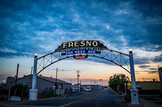 An illuminated sign spans across a road, reading 'Fresno, the best little city in the U.S.A.' with 'Van Ness Ave' in blue neon underneath. The photo is taken during twilight featuring a partly cloudy sky with shades of blue and orange, and the street is calm with minimal traffic and industrial buildings on either side.