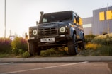 A rugged, matte black Land Rover Defender is parked on a grassy area near a curb. The headlights are on, and it's positioned against a backdrop of an industrial building with orange accents. Wildflowers and greenery surround the vehicle, and the lighting suggests it's about dusk.