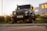 A rugged, matte black Land Rover Defender is parked on a grassy area near a curb. The headlights are on, and it's positioned against a backdrop of an industrial building with orange accents. Wildflowers and greenery surround the vehicle, and the lighting suggests it's about dusk.