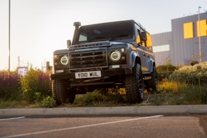 A rugged, matte black Land Rover Defender is parked on a grassy area near a curb. The headlights are on, and it's positioned against a backdrop of an industrial building with orange accents. Wildflowers and greenery surround the vehicle, and the lighting suggests it's about dusk.