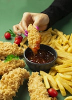 A hand with colorful nails is dipping a piece of crispy fried chicken into a bowl of dark, spicy sauce. The table is filled with golden French fries, additional pieces of chicken, and red cherry tomatoes. Green parsley leaves add a touch of color to the arrangement.
