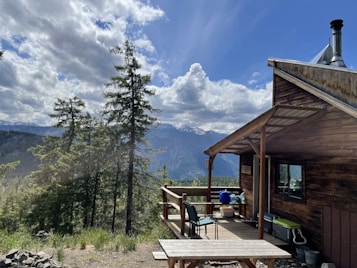 A wooden cabin with a small porch is set amidst a mountainous landscape. Tall pine trees surround the cabin, and in the background, there are snow-capped mountains under a partly cloudy sky. The porch has a chair and a small table with outdoor items.