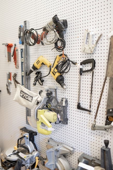 A white pegboard with various tools neatly arranged, including power drills, saws, wiring, and clamps. The tools have different colors and shapes, and the board allows for organized storage.
