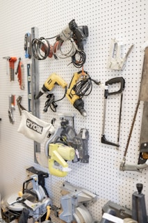 A white pegboard with various tools neatly arranged, including power drills, saws, wiring, and clamps. The tools have different colors and shapes, and the board allows for organized storage.