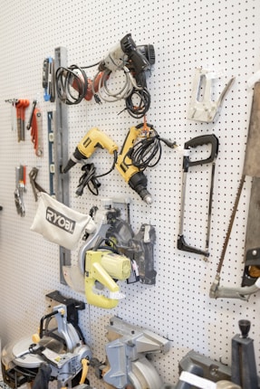 A white pegboard with various tools neatly arranged, including power drills, saws, wiring, and clamps. The tools have different colors and shapes, and the board allows for organized storage.
