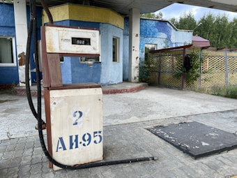 An old, seemingly abandoned gas station with a rusted fuel pump displaying 'AI-95' and the number 2. The background includes a weathered blue and yellow building with visible peeling paint. There are tall trees partially visible, and a lattice fence with greenery surrounding the area.