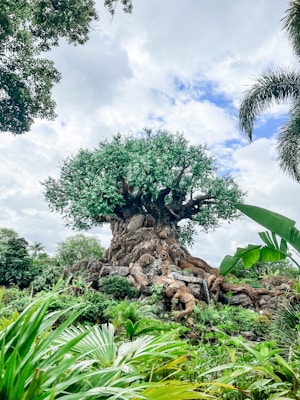 A massive, intricately detailed artificial tree stands prominently in a lush, green landscape. Its trunk is artistically sculpted with various animal carvings, giving it a mythical appearance. The foliage is dense and vibrant, surrounded by diverse tropical plants.