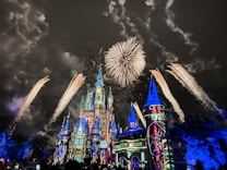 A brightly illuminated castle with intricate architectural details is the centerpiece, surrounded by a nighttime sky filled with vibrant, golden fireworks. The castle features colorful lighting projections, giving it a magical appearance.